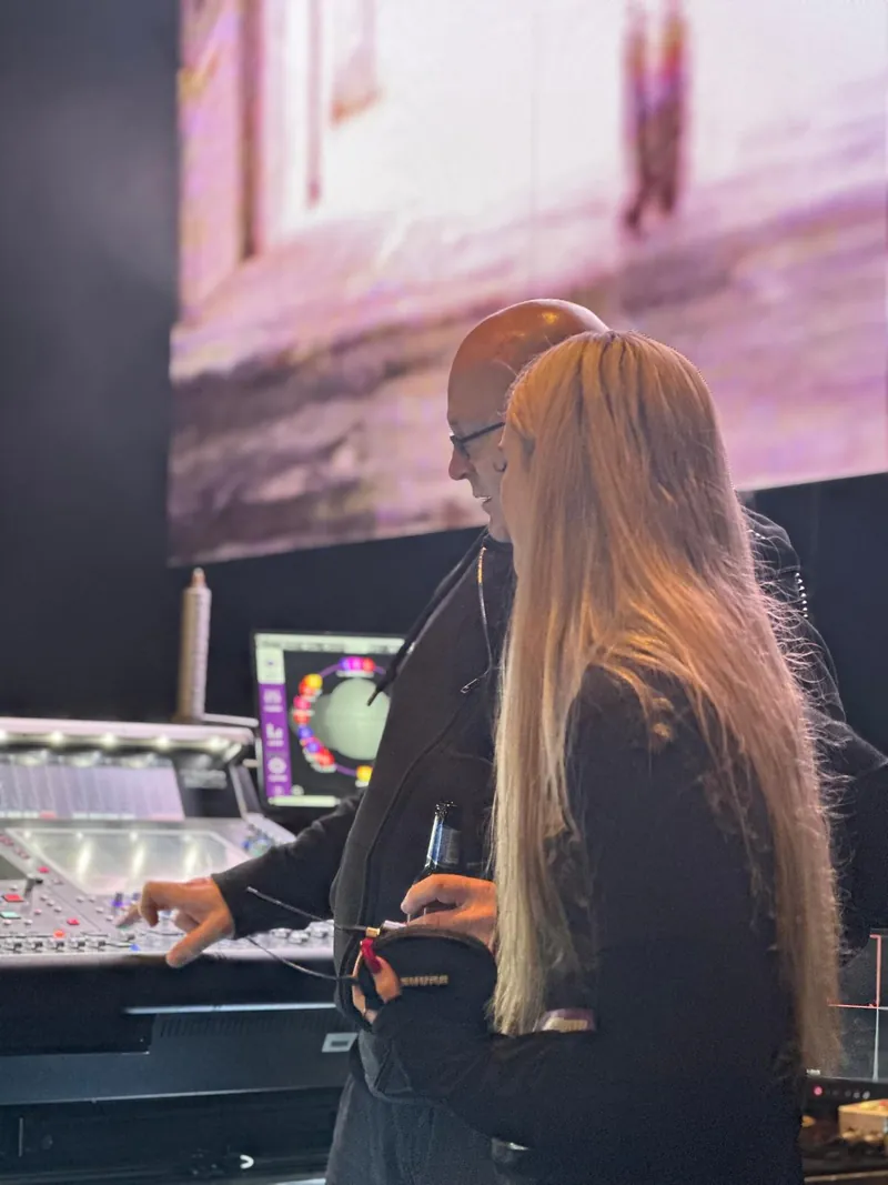 Mike Osman at a DiGiCo console talking through the mix with a woman holding a Shure headset microphone, in front of a large video screen